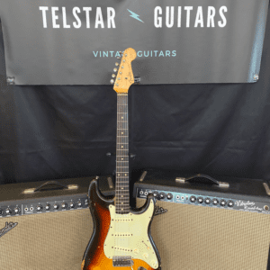 The image shows a vintage sunburst electric guitar on a stand, positioned between two vintage-style amplifiers. The guitar features a light wood neck and a classic sunburst finish on the body. In the background is a black banner with the text "Telstar Guitars Vintage Guitars ESTD 2019." The scene is set on a patterned rug, creating a vintage, retro ambiance.