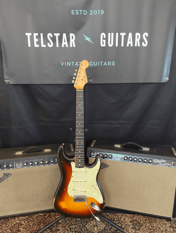 The image shows a vintage sunburst electric guitar on a stand, positioned between two vintage-style amplifiers. The guitar features a light wood neck and a classic sunburst finish on the body. In the background is a black banner with the text "Telstar Guitars Vintage Guitars ESTD 2019." The scene is set on a patterned rug, creating a vintage, retro ambiance.