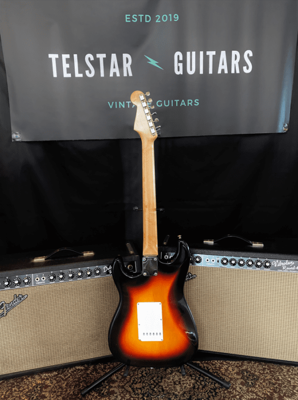 The image shows the back view of a vintage sunburst electric guitar standing on a display between two vintage-style amplifiers. The guitar has a wooden neck, and the body has a sunburst finish. Behind the guitar is a black banner with the text "Telstar Guitars Vintage Guitars ESTD 2019." The guitar and amplifiers are set on a patterned rug, creating a classic, vintage atmosphere.