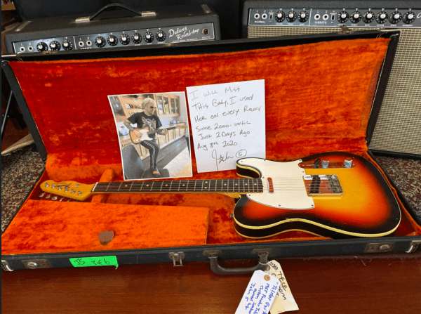 The image shows a sunburst electric guitar placed inside an open, orange-lined guitar case. In the case, there is a handwritten note that reads, "I will miss this baby. I used her on every record since 2000 until just 2 days ago, Aug 7th, 2020 - John 5," along with a photograph of a person playing the guitar. In the background, Fender amplifiers are visible. The guitar has a white pickguard and is accompanied by a label with additional handwritten details attached to the case.