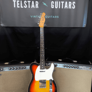 The image shows a vintage-style electric guitar with a sunburst finish and a white pickguard, displayed in front of two Fender amplifiers. The guitar is upright, with the front of the body and fretboard visible. Behind the guitar is a banner that reads “Telstar Guitars” with “Vintage Guitars” and "ESTD 2019" above it. The amplifiers and patterned rug add to the vintage aesthetic of the setup.