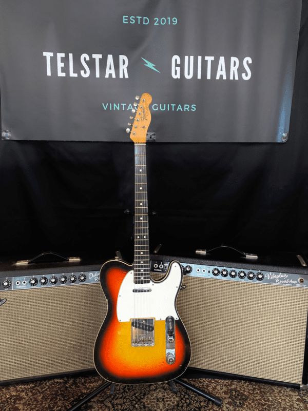 The image shows a vintage-style electric guitar with a sunburst finish and a white pickguard, displayed in front of two Fender amplifiers. The guitar is upright, with the front of the body and fretboard visible. Behind the guitar is a banner that reads “Telstar Guitars” with “Vintage Guitars” and "ESTD 2019" above it. The amplifiers and patterned rug add to the vintage aesthetic of the setup.