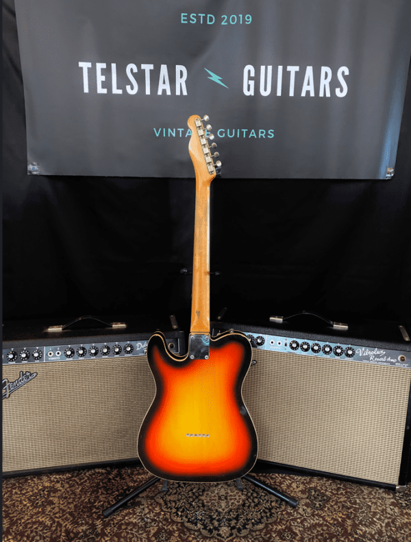 The image depicts a sunburst electric guitar, displayed standing vertically in front of two Fender amplifiers. The guitar is positioned with its back facing forward, showing the wooden neck and body. Behind the guitar is a banner that reads "Telstar Guitars" with "Vintage Guitars" written below it, along with the text "ESTD 2019." The guitar and amplifiers rest on a patterned rug, contributing to the vintage aesthetic of the setup.