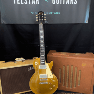 The image shows a gold Gibson Les Paul electric guitar standing upright between two vintage Gibson amplifiers. The setup is placed on a patterned rug, and a banner behind the guitar reads "Telstar Guitars," with "Vintage Guitars" and "ESTD 2019" displayed above it. The amplifiers, guitar, and banner together create a vintage, classic guitar display.