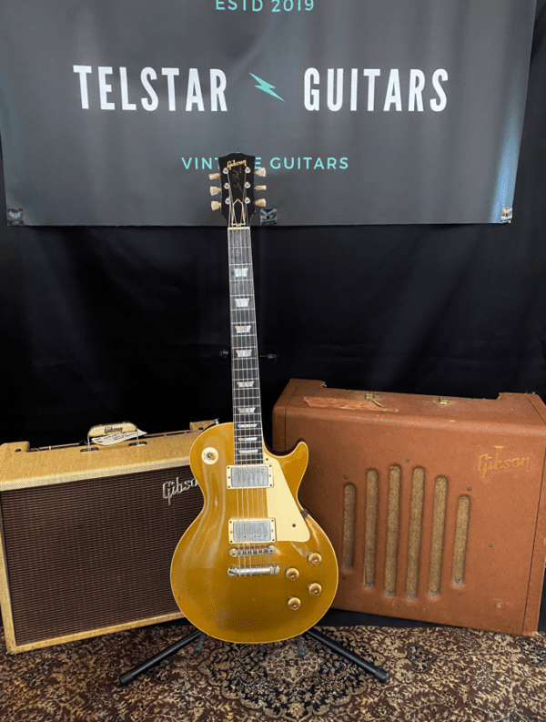 The image shows a gold Gibson Les Paul electric guitar standing upright between two vintage Gibson amplifiers. The setup is placed on a patterned rug, and a banner behind the guitar reads "Telstar Guitars," with "Vintage Guitars" and "ESTD 2019" displayed above it. The amplifiers, guitar, and banner together create a vintage, classic guitar display.