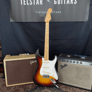 The image shows the front view of a vintage Fender Stratocaster guitar with a sunburst finish and white pickguard, displayed on a stand. The guitar is positioned between a Gibson amplifier on the left and a Fender amplifier on the right. The setup is placed on a patterned rug, with a black banner in the background that reads "Telstar Guitars Vintage Guitars ESTD 2019." The image highlights the classic look and design of the guitar along with the vintage-style amplifiers.